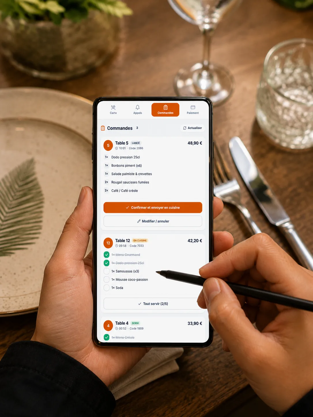 Une serveuse coche les commandes au stylet sur la page de contrôle Winacom affichée sur son smartphone (suivi Confirmé → En cuisine → Servi)